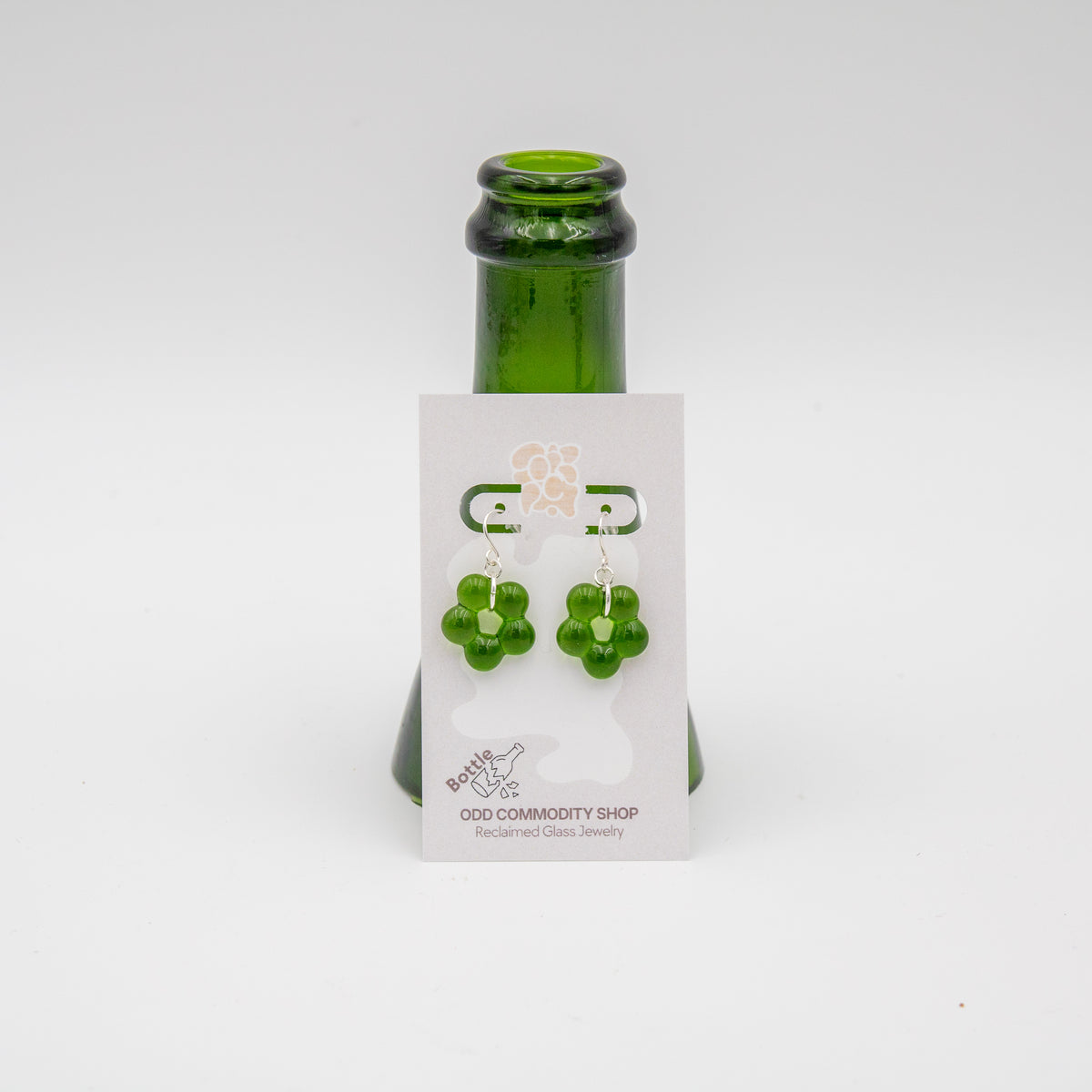 Dangly flower shaped green pair of earrings on a branded card leaning against a cut wine bottle top. 