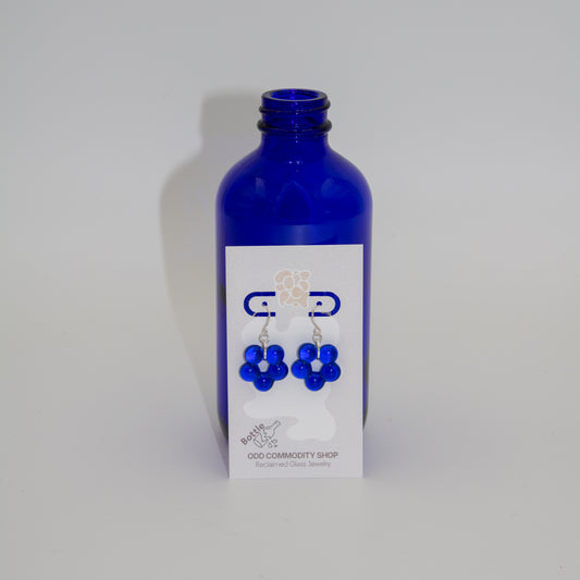 A pair of dangle earrings featuring five translucent cobalt blue beads arranged in a flower-like cluster, suspended from silver fishhook wires, shown on a neutral light gray background with blue bottle behind the product card.