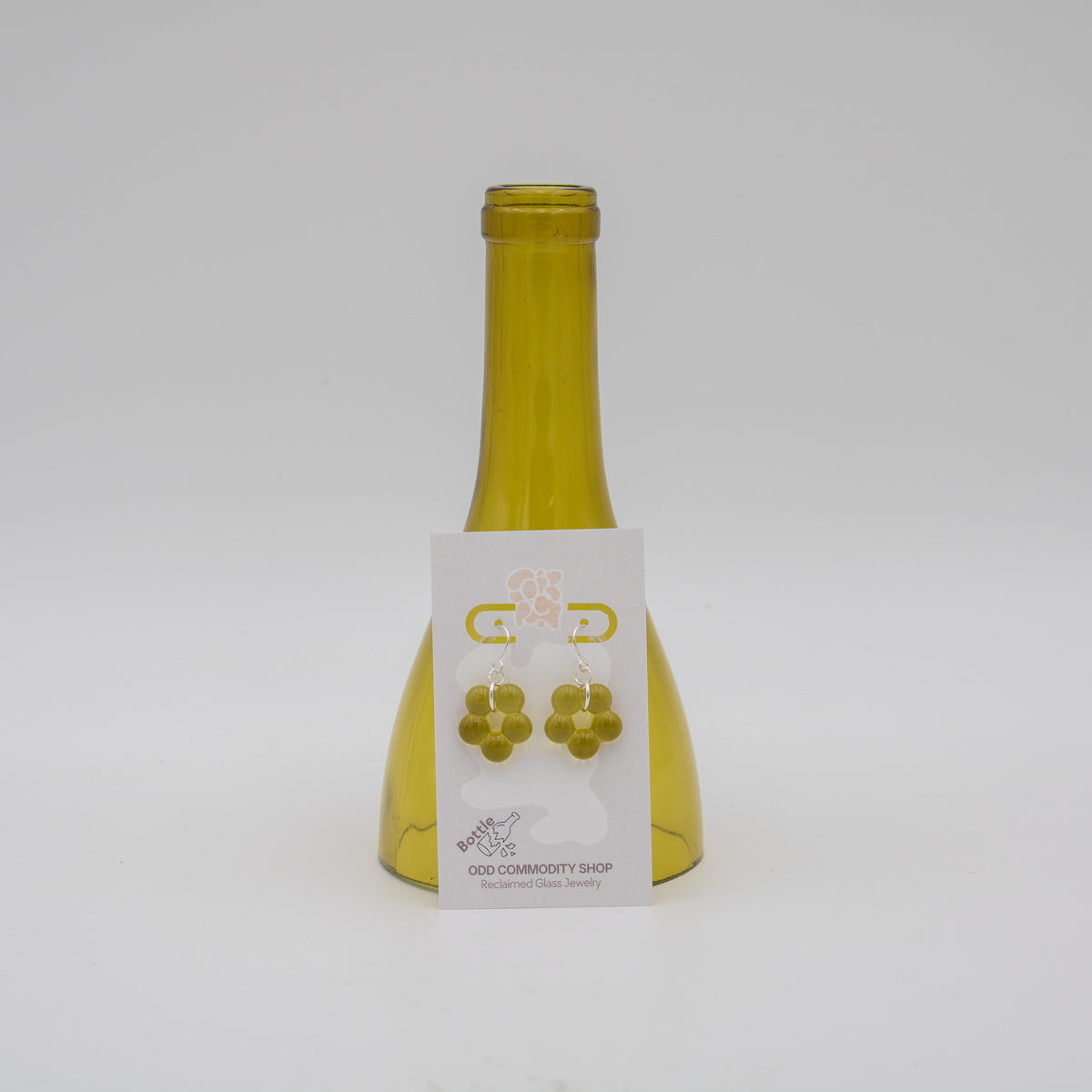 A pair of gold flower-shaped stud earrings displayed on a white backing card, which is attached to a translucent olive-green glass bottle. The product is photographed against a seamless light gray background.