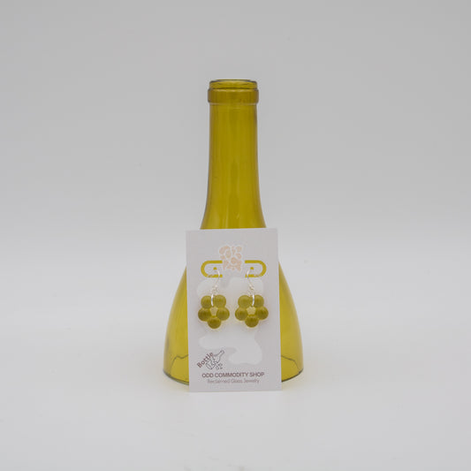 A pair of gold flower-shaped stud earrings displayed on a white backing card, which is attached to a translucent olive-green glass bottle. The product is photographed against a seamless light gray background.