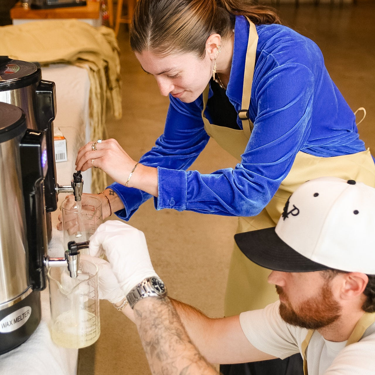 Person in a blue shirt pouring liquid from a coffee machine into a container, with another person in a white shirt and black cap assisting.