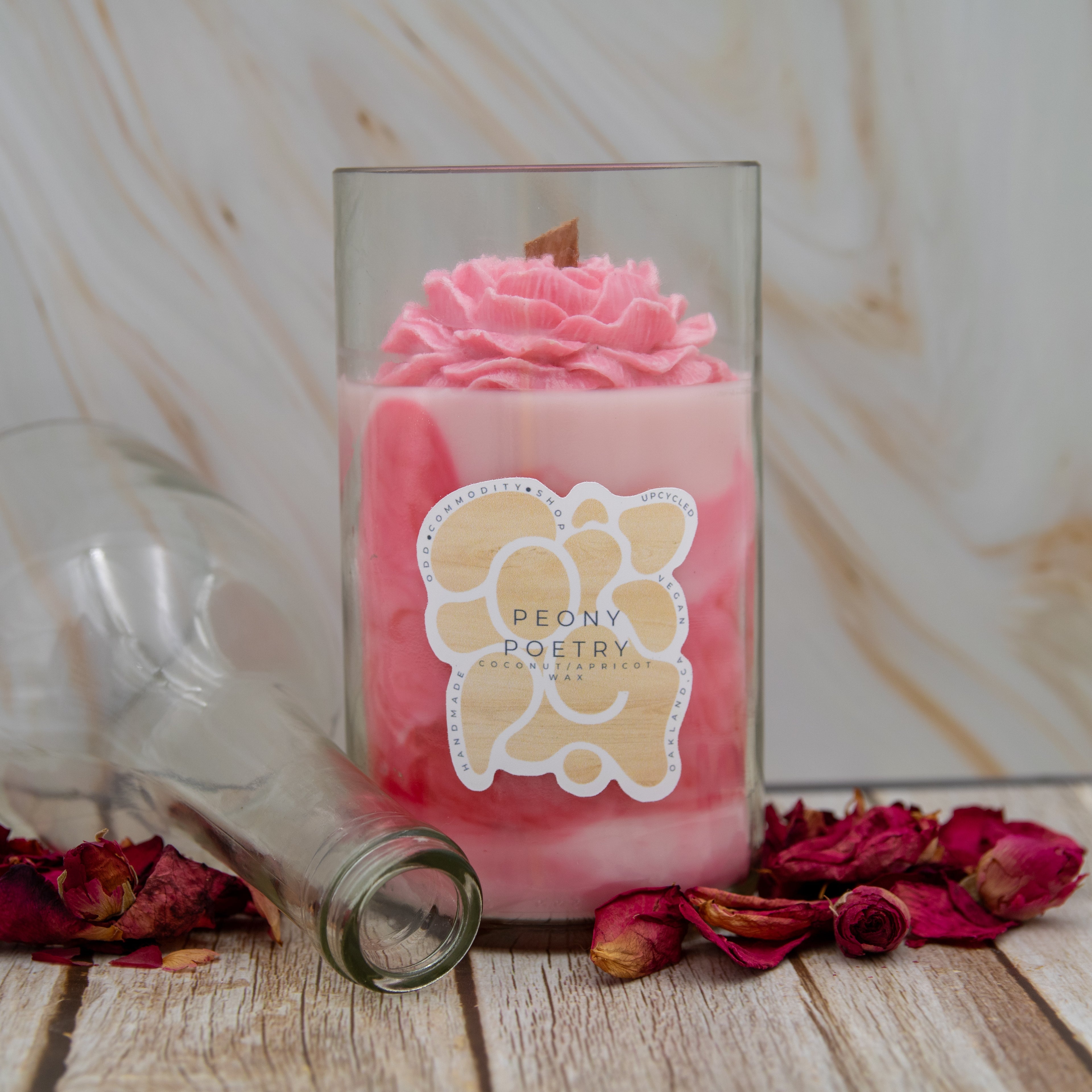Pink candle in a glass jar with a 'Peony Poetry' label, surrounded by dried roses on a wooden surface.