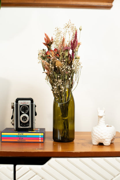 Decorative setup with a vintage camera, books, and an upcycled wine bottle vase with flowers on a wooden surface.