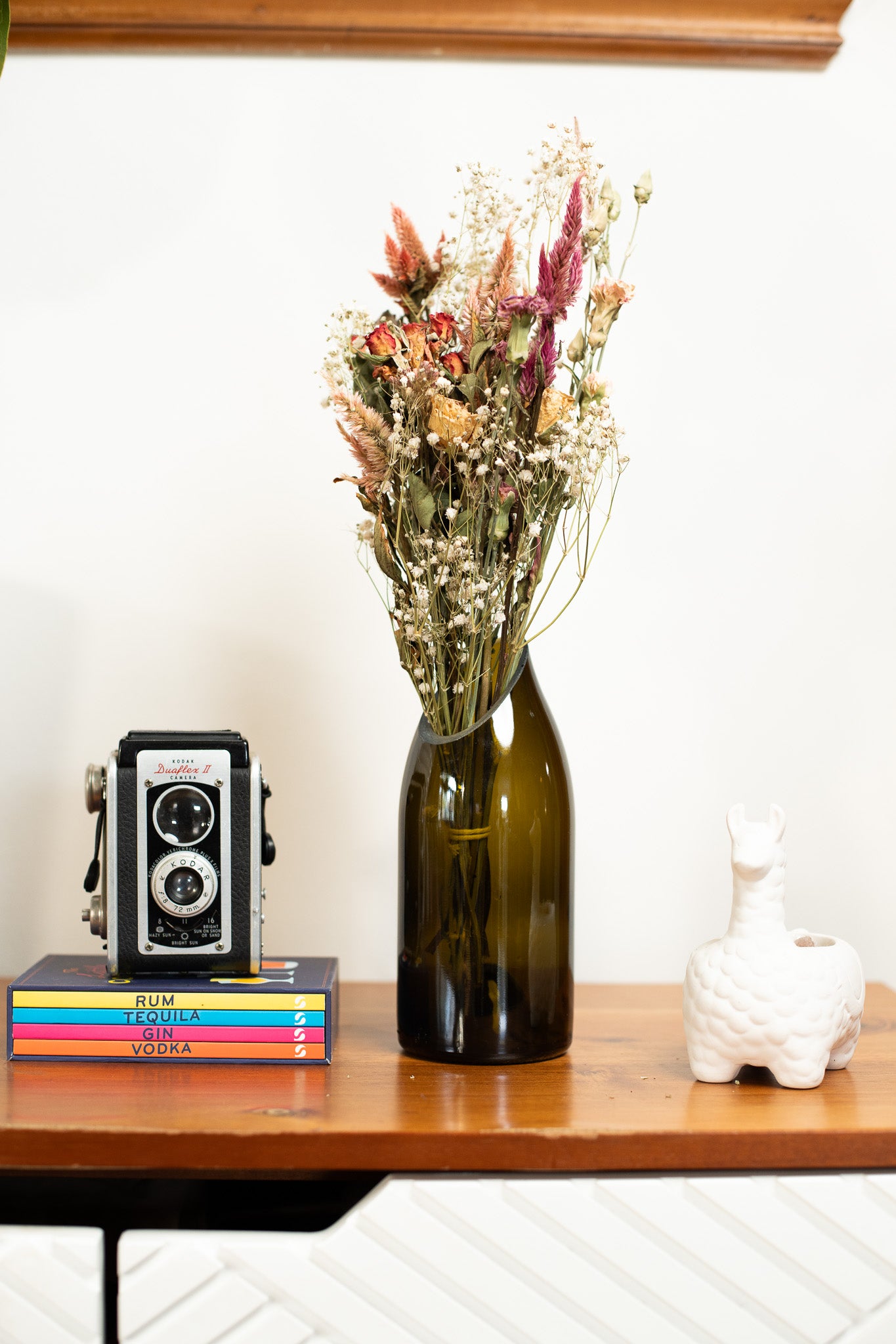 Decorative setup with a green bottle, dried flowers, vintage camera, and books on a wooden surface.
