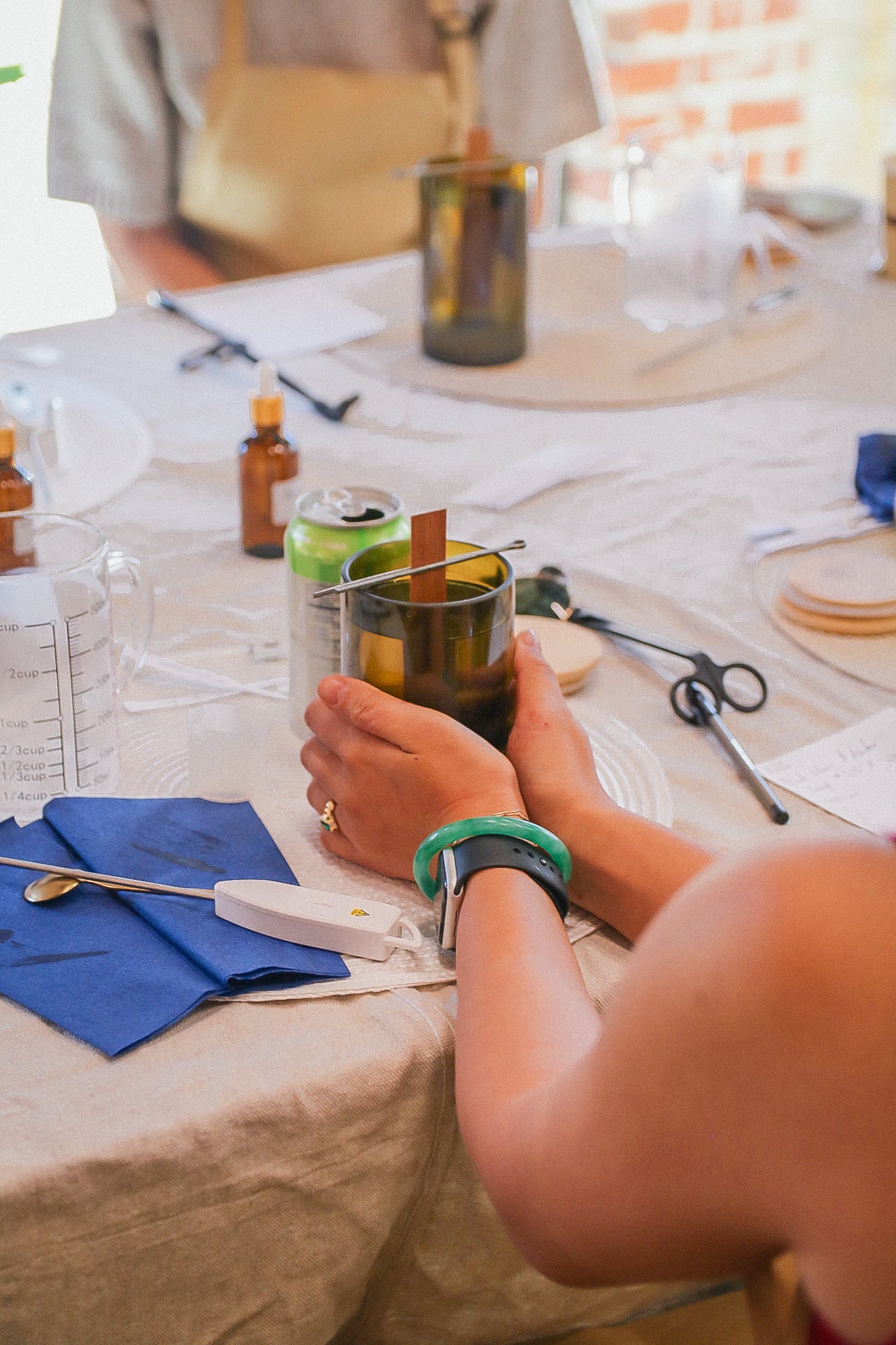 Person sitting at a table holding reclaimed glass candle surrounded by various items and candle making materials including thermometers, beakers, and snacks. 