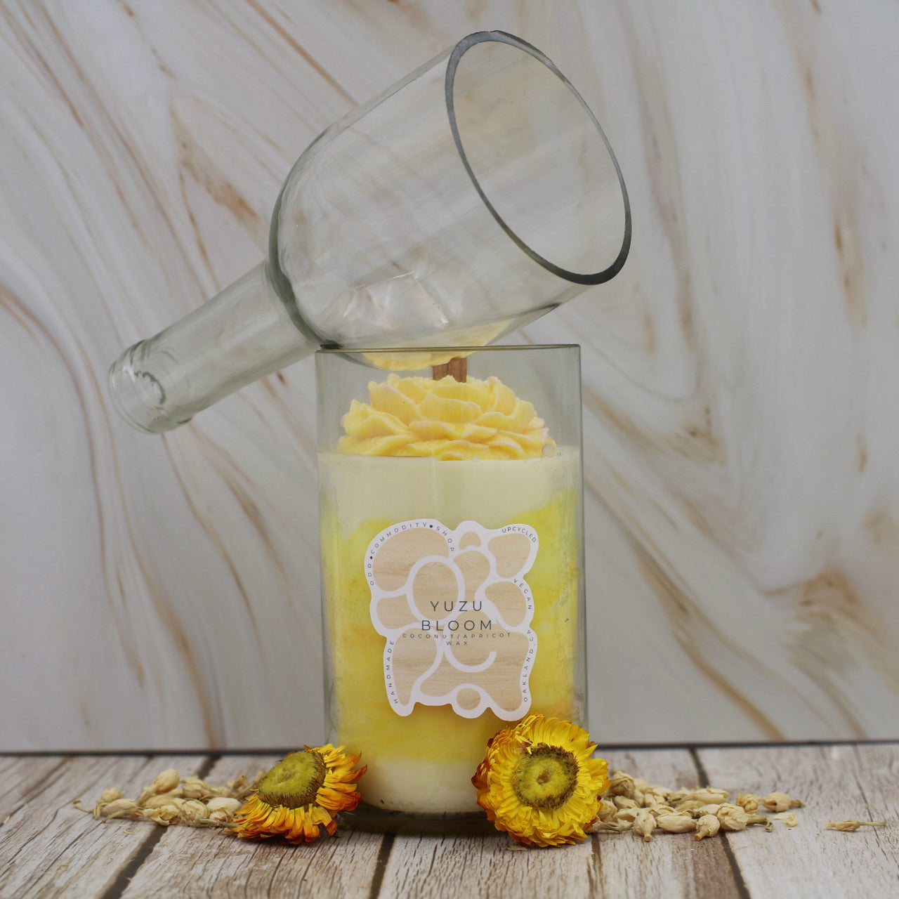Candle made from upcycled wine bottle with a floral design and 'Yuzu Bloom' label, surrounded by dried flowers on a wooden surface.