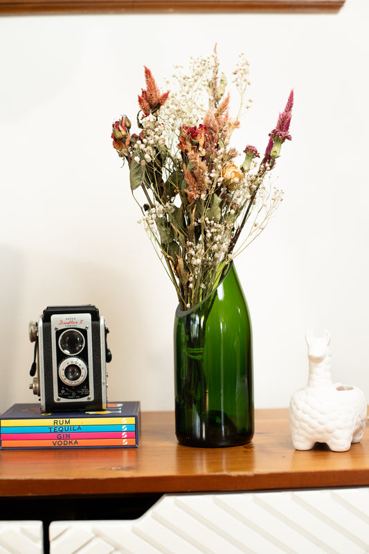 Green vase with dried flowers on a wooden shelf with books and a camera.