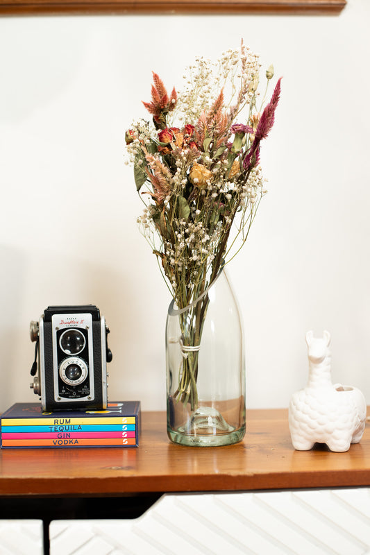 Vase with dried flowers on a wooden surface next to books and a camera.