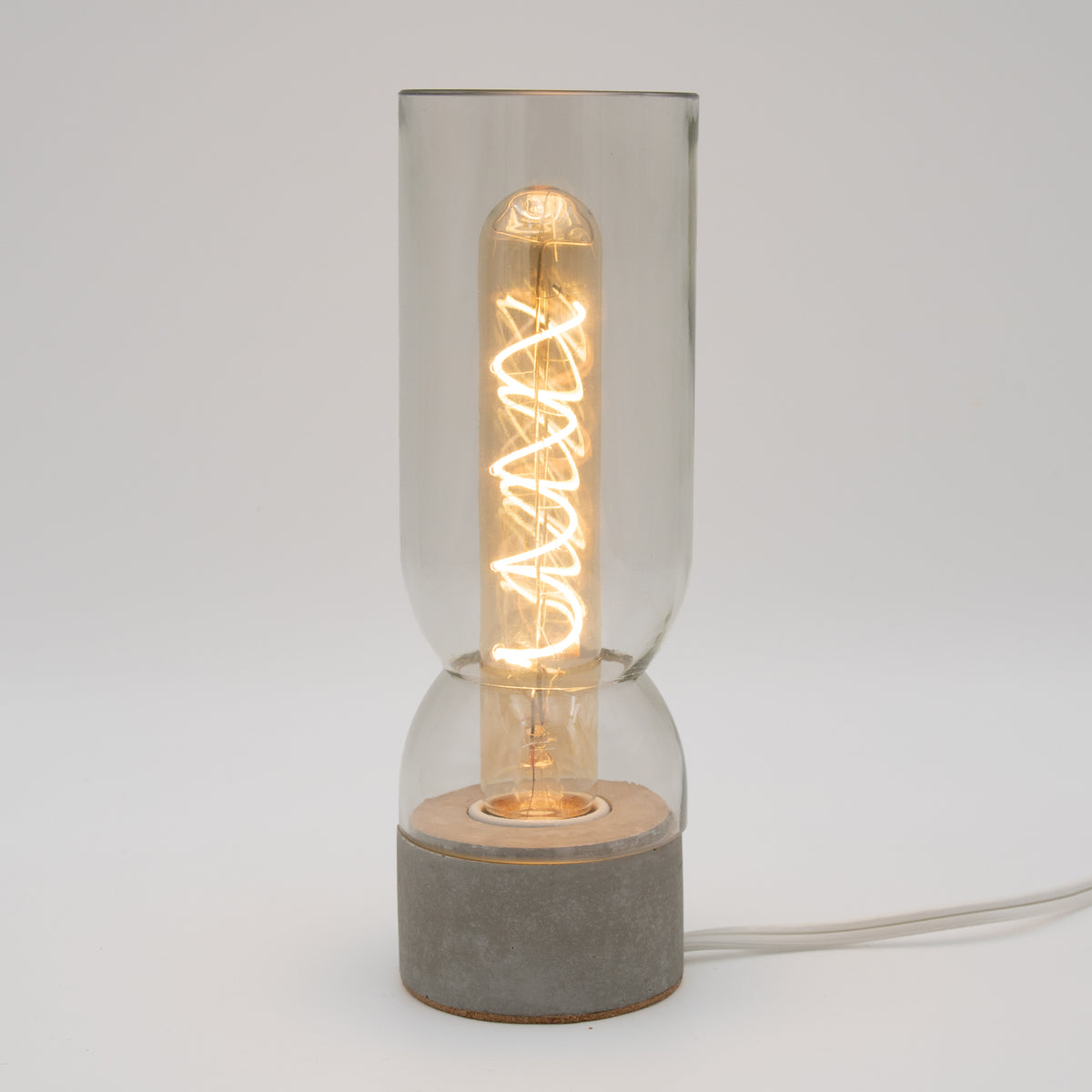 A modern table lamp with a glowing warm white spiral filament bulb inside a clear glass cylinder, set on a round concrete base with a light wood top, shown against a neutral light gray background.