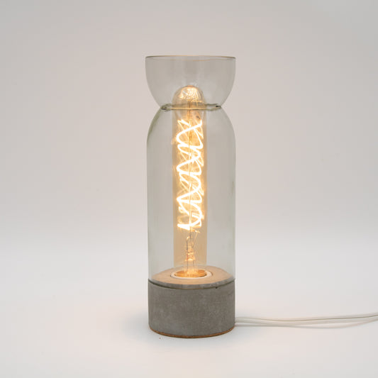 A modern industrial-style table lamp with a glowing spiral filament bulb inside a clear glass cylinder, set on a round concrete base against a neutral light gray background.